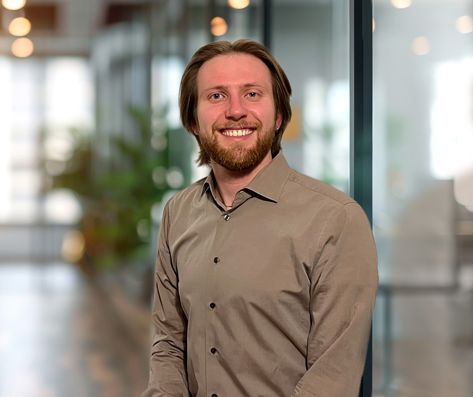 A smiling man with shoulder-length light brown hair and a beard wears a taupe button-down shirt in an office.