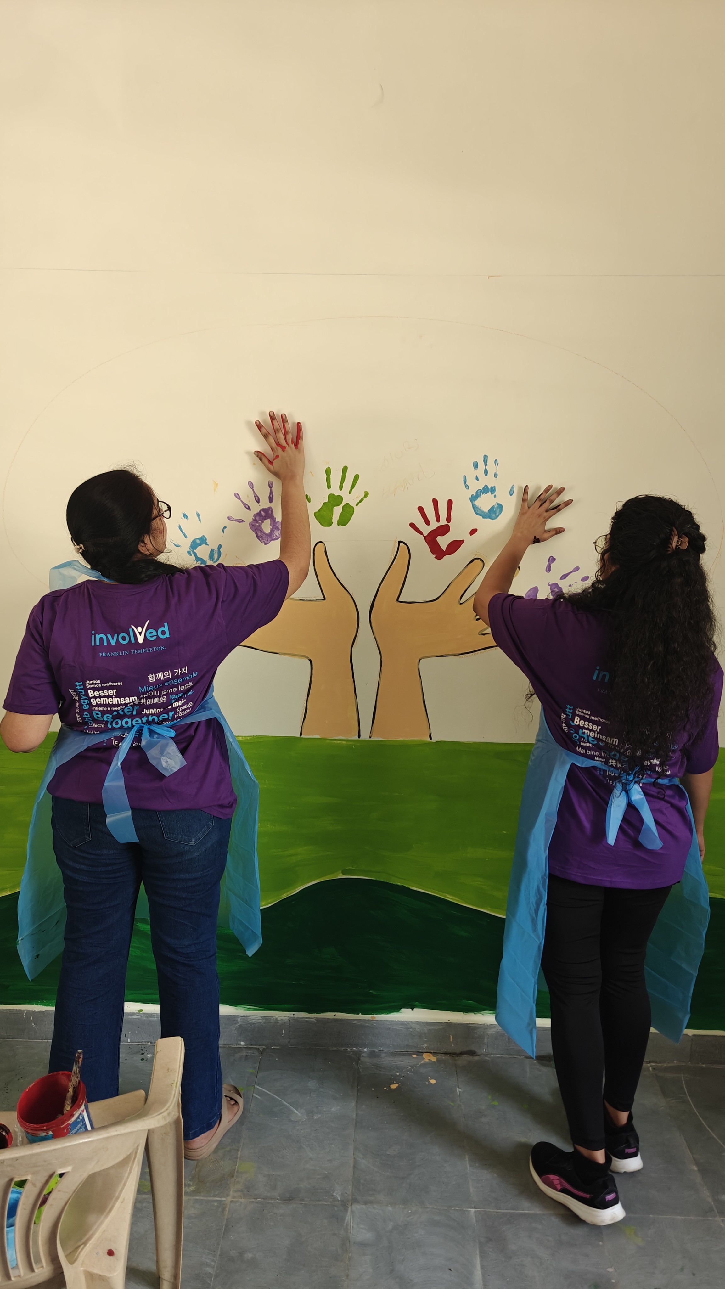 Two women painting a mural on a wall, featuring three large brown hands reaching upward with colorful handprints in purple, green, red, and blue above them.