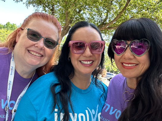 Three women outdoors, smiling and posing closely for a selfie, all wearing shirts with text and sunglasses.