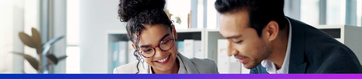 A woman with dark curly hair and glasses smiles while sitting at a desk in a modern office. A male colleague in a suit stands beside her, holding a tablet and pointing at the screen as they collaborate.