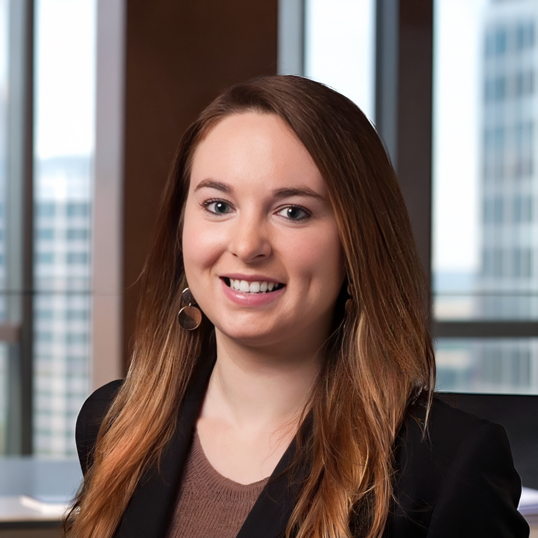 Headshot of a smiling woman with long light brown hair, wearing a black blazer and brown top, in an office.