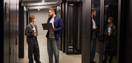 Two IT technicians, a man and a woman, stand in a brightly lit hallway of a data center, flanked by rows of tall black server racks.