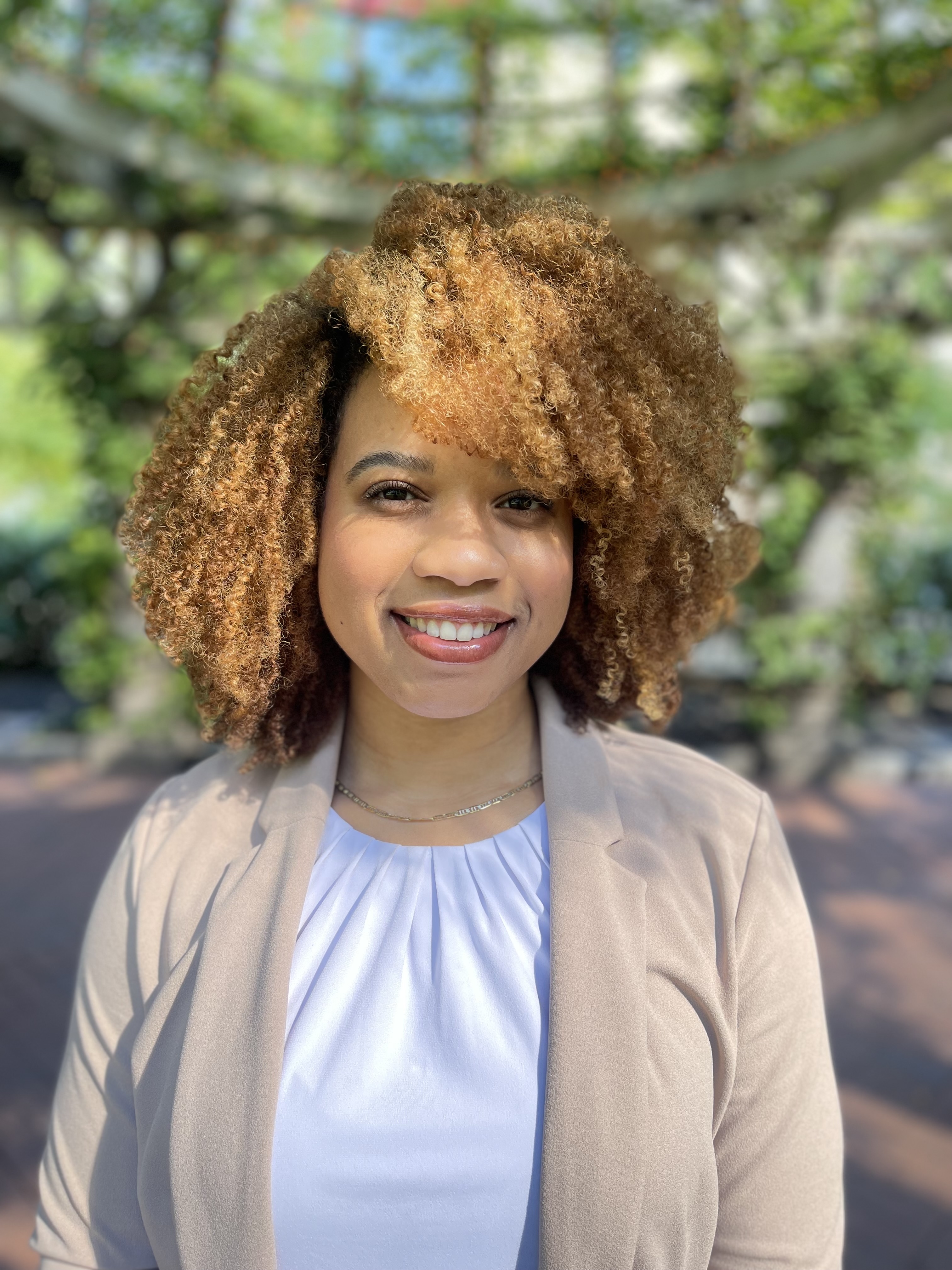 A smiling woman with curly, honey-blonde hair poses outdoors for a headshot. She wears a white pleated blouse, tan blazer, and gold necklace, with a blurred green garden in the background.