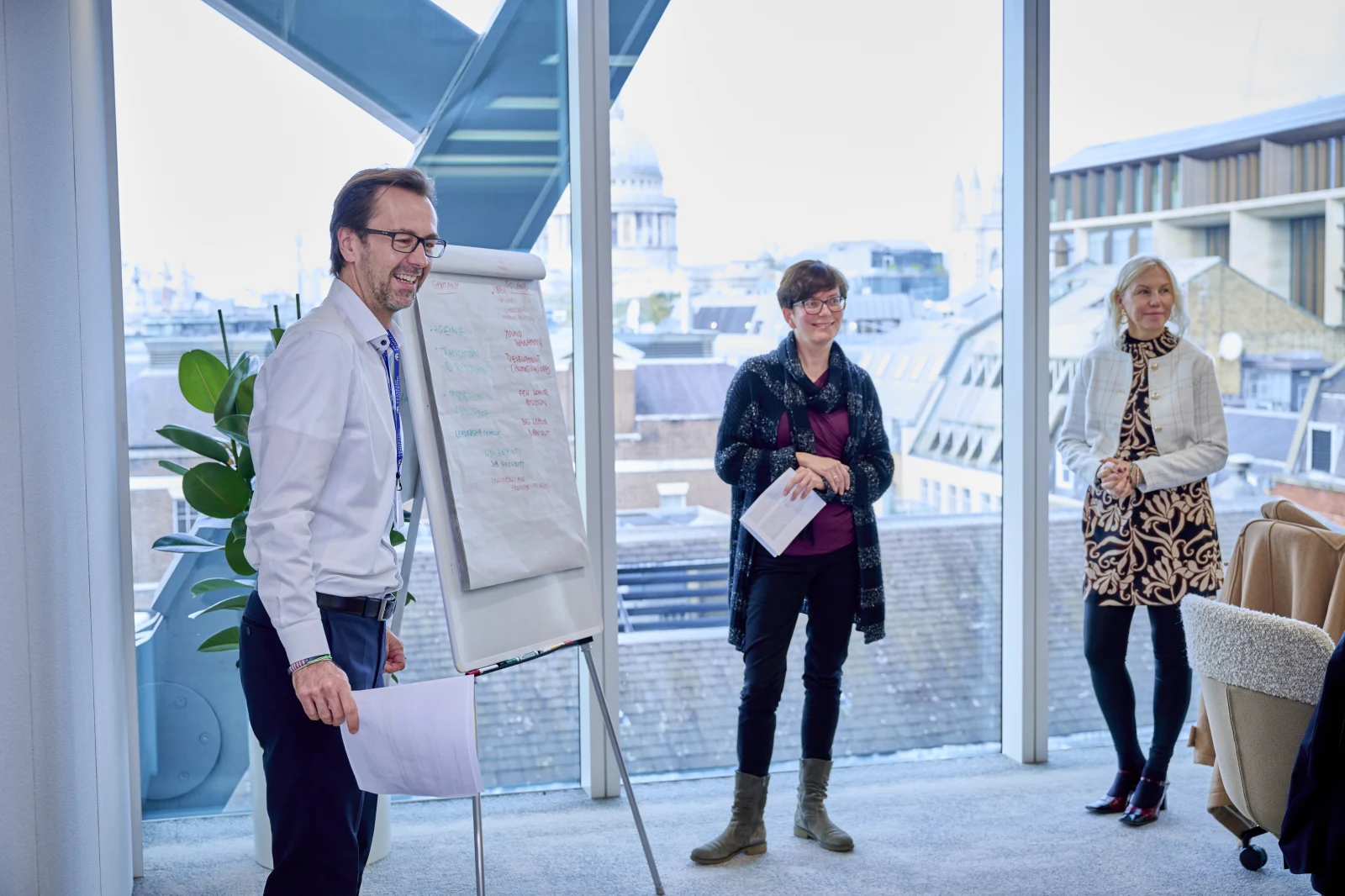 Three businesspeople standing in a meeting room, one presenting to the others near a whiteboard. Large windows show a city skyline.