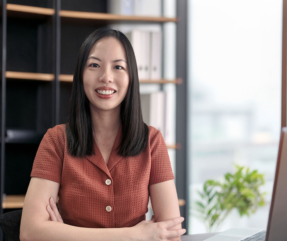 Professional headshot of woman with long, dark hair, wearing a burnt-orange, short-sleeved collared shirt, and sitting at a desk in a modern office