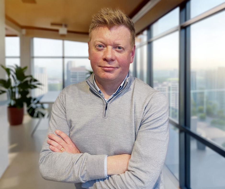 Professional headshot of a man with short blond hair and blue eyes. He is wearing a grey quarter-zip sweater over a collared shirt and stands with his arms crossed in a modern office