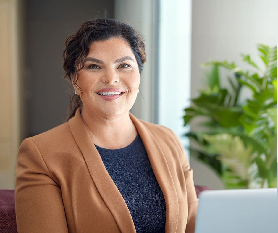 Professional headshot of a woman with dark wavy hair pulled back, wearing a tan blazer over a dark blue top, smiling warmly at the camera in a bright, modern office setting.