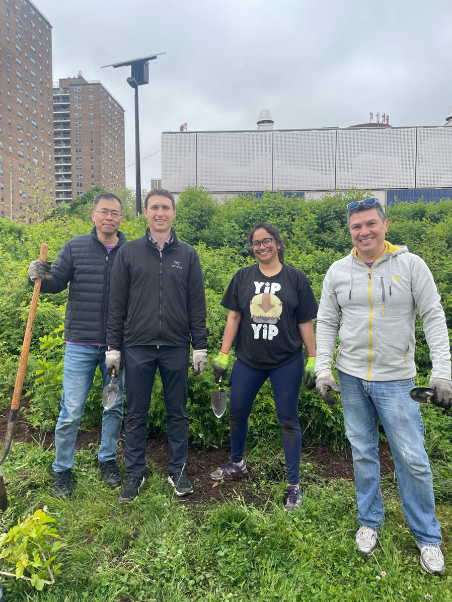 Three men and one woman stand on a grassy hill, posing for a photo. One man holds a shovel. Tall buildings are visible in the background.