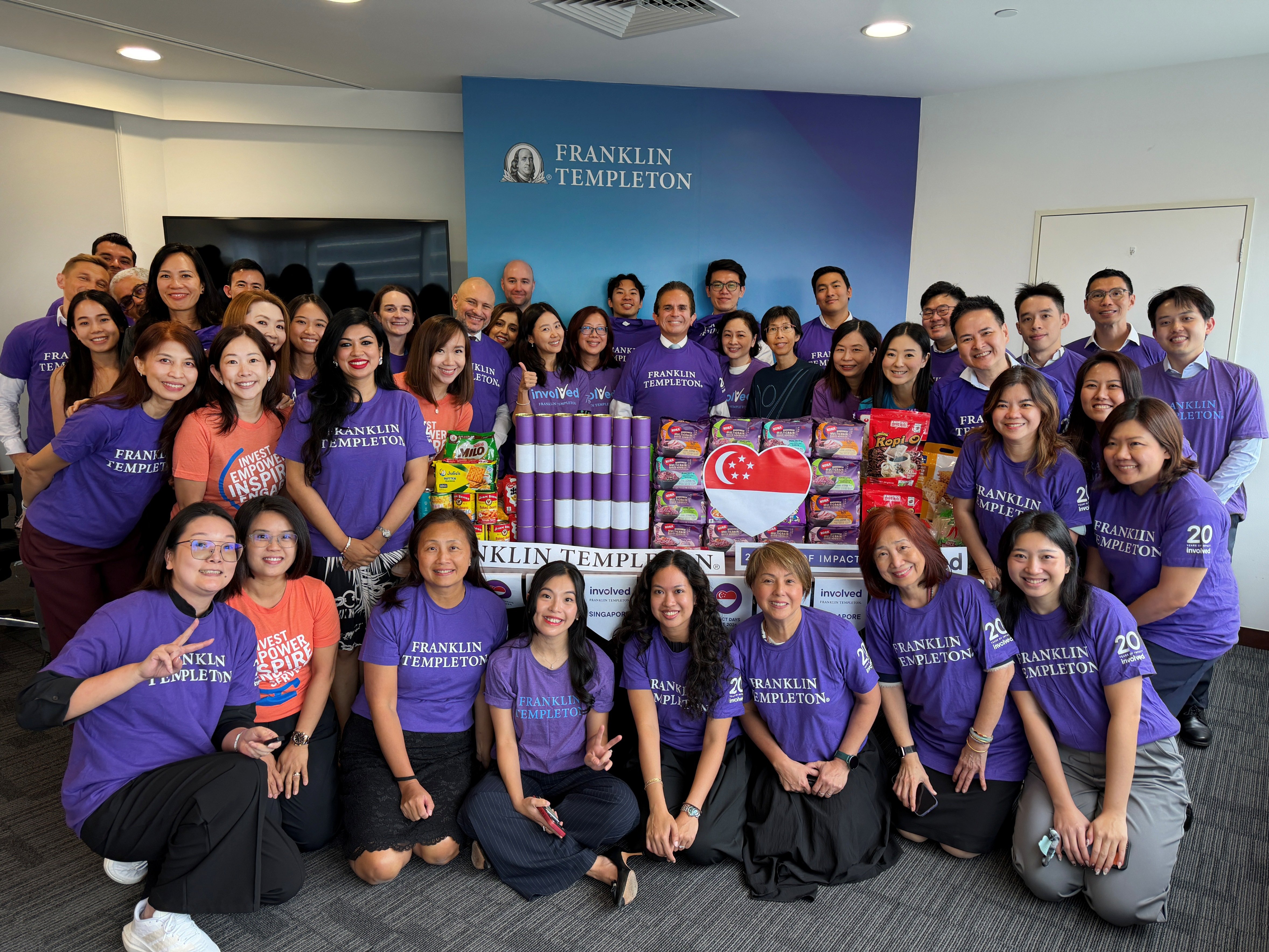 A large group of Franklin Templeton employees, most wearing matching purple shirts, pose in an office around a table filled with donated food items.