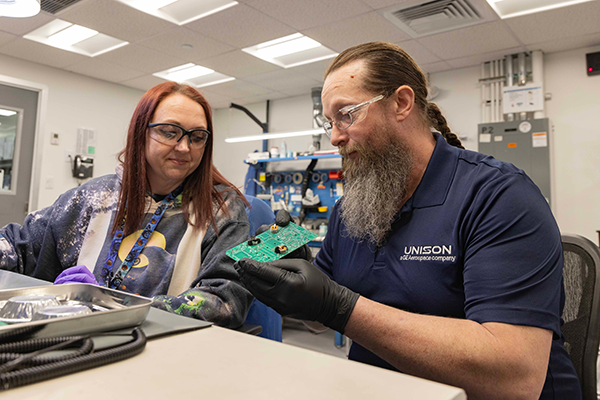 Two people in an industrial setting examine a green circuit board, with tools and equipment visible in the background.