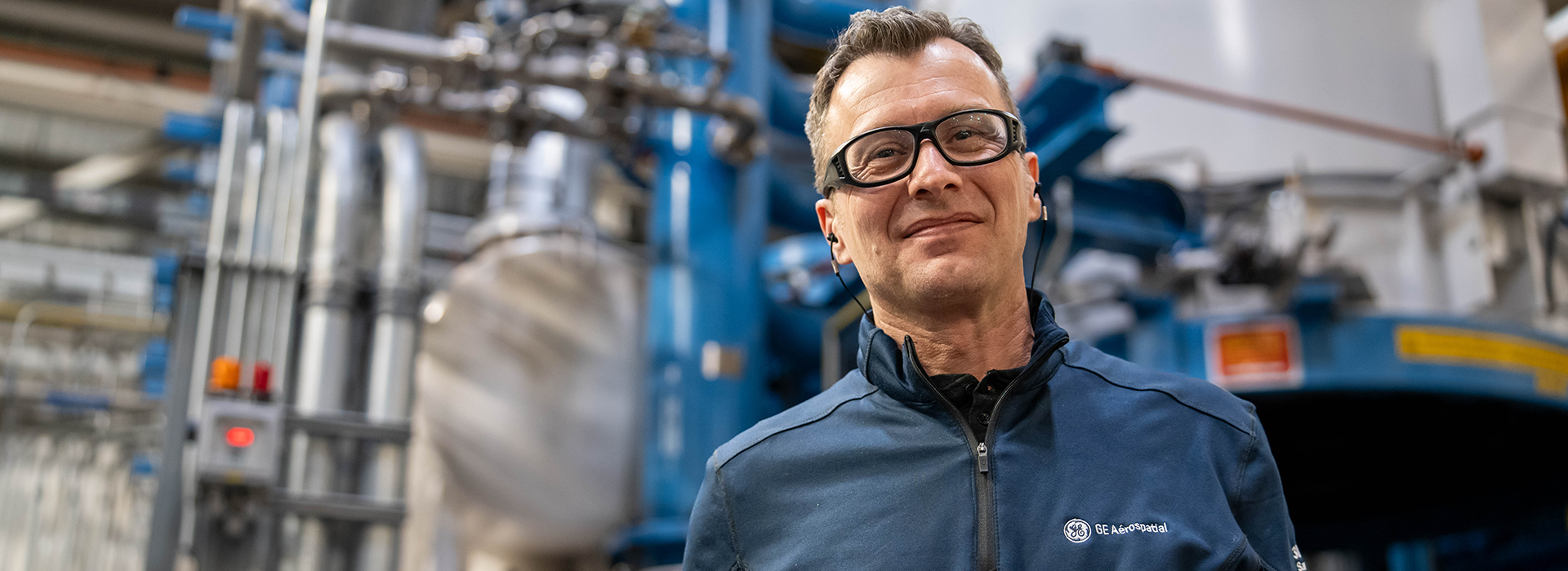 A person wearing a navy blue GE Aerospace jacket stands in front of industrial equipment, including pipes, machinery, and control panels in a manufacturing facility.