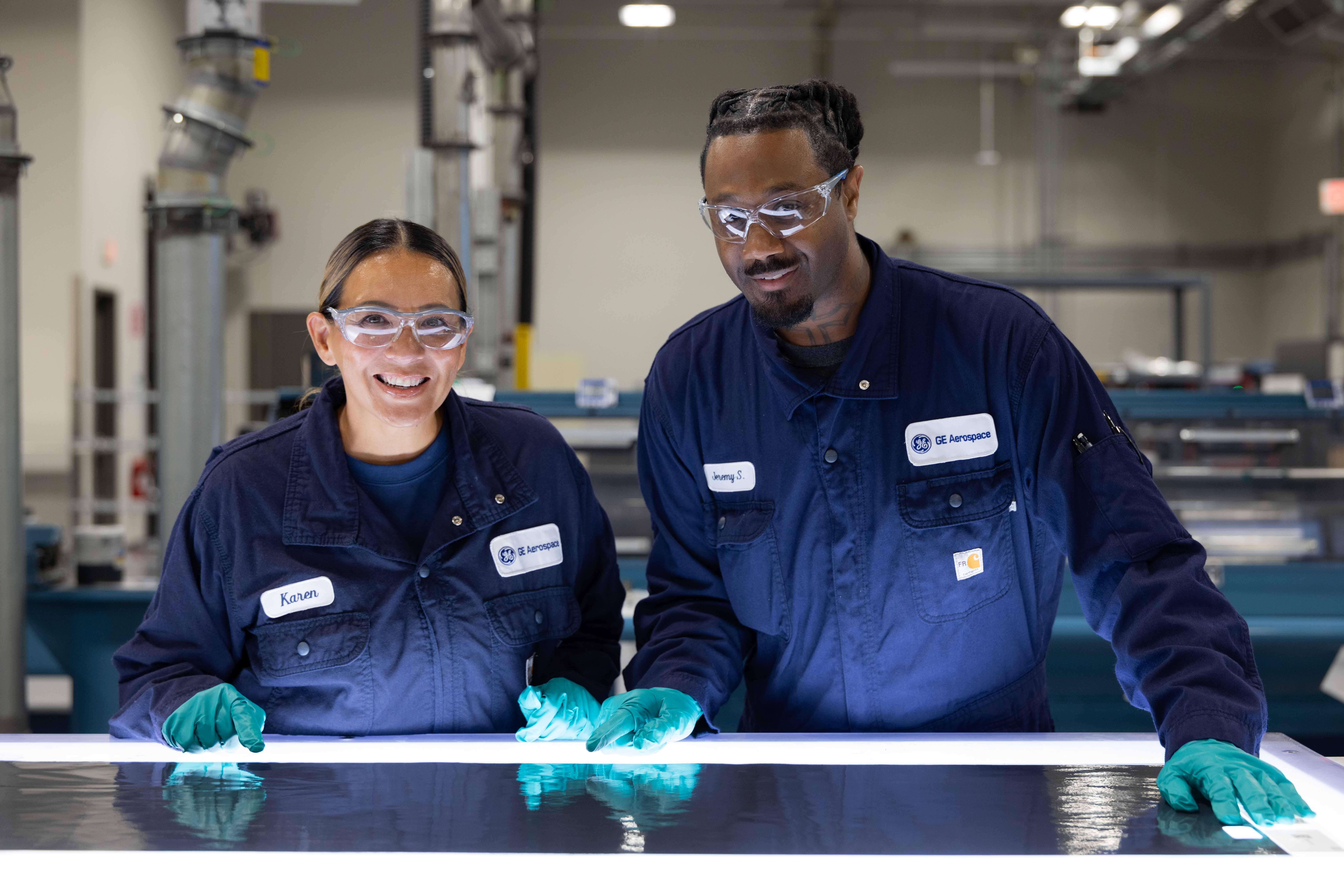 Two GE Aerospace employees wearing navy blue uniforms and green gloves are working together at a brightly lit workstation. The workstation features a glowing surface, and the employees appear to be inspecting or handling a flat material. The background includes industrial equipment and a manufacturing environment.