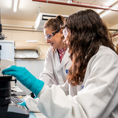 Two individuals in lab coats and safety gloves work together at a microscope in a laboratory setting.