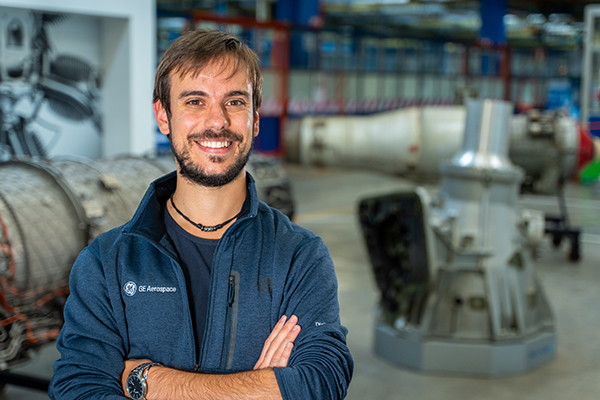 Man smiling at camera with arms crossed in a factory setting.