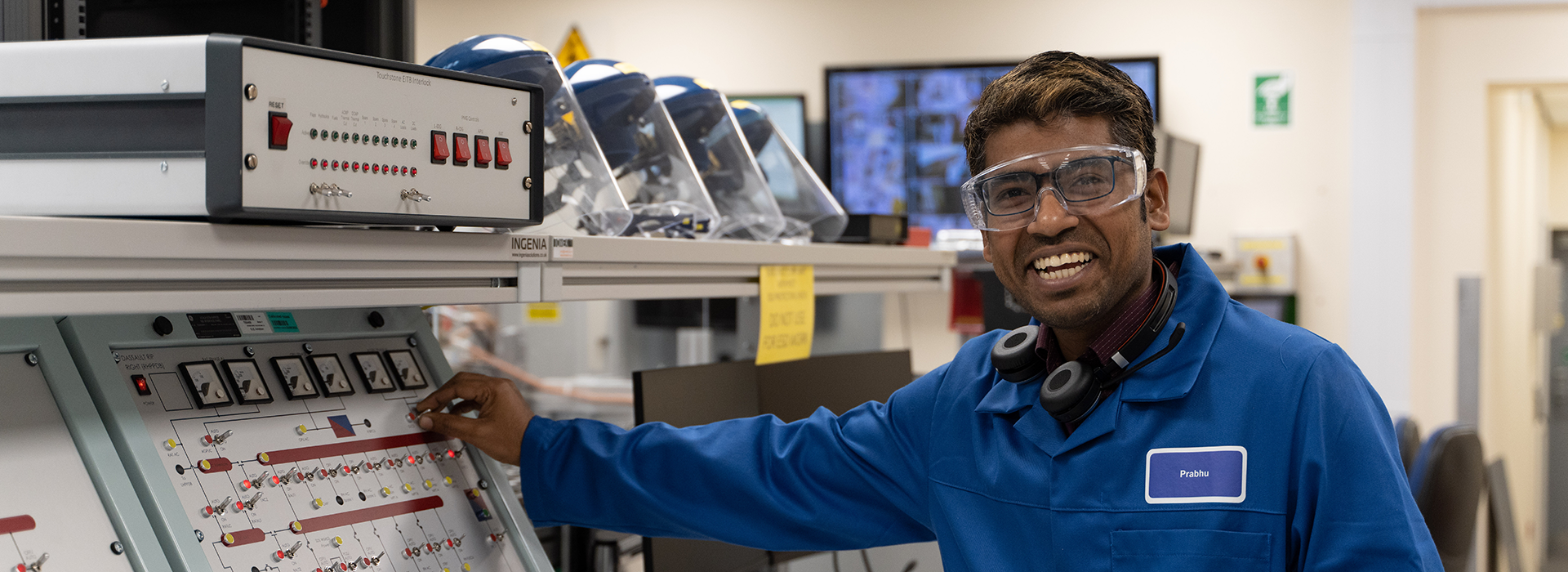 A person wearing a blue lab coat is working in a technical lab environment. They are interacting with a control panel featuring various switches, buttons, and indicator lights. In the background, there are protective helmets with visors, monitors displaying images, and other lab equipment. The setting appears to be a professional workspace focused on engineering or testing.