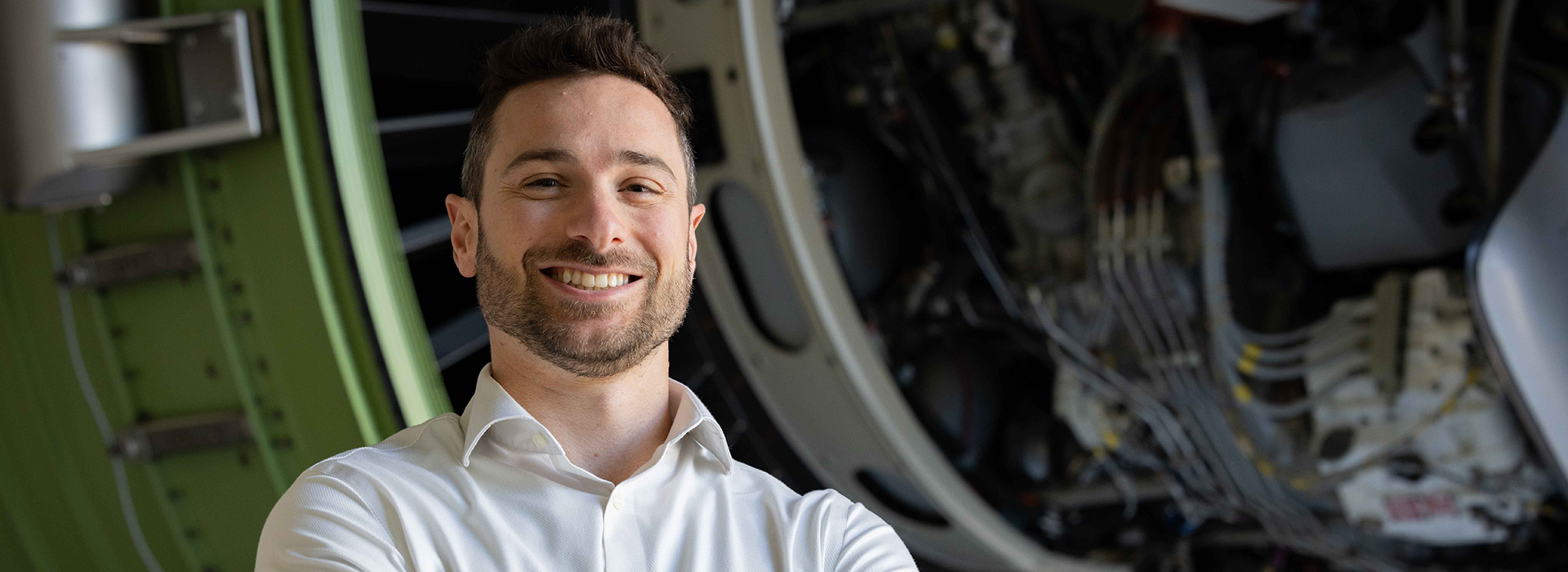 A person wearing a white collared shirt stands in front of an aircraft engine assembly, showcasing intricate wiring, components, and a green structural frame.