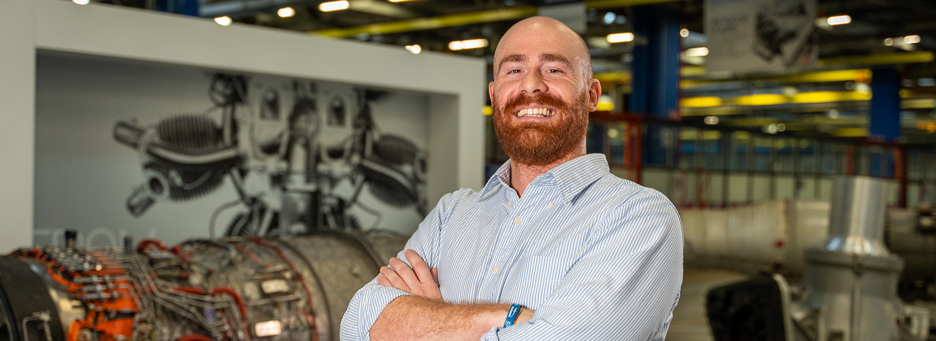 A person wearing a light blue striped button-down shirt stands with arms crossed in an industrial setting. Behind them, there is a large engine component with visible wiring and machinery, as well as a wall featuring a detailed technical illustration of engine parts. The background includes yellow beams, equipment, and signage.