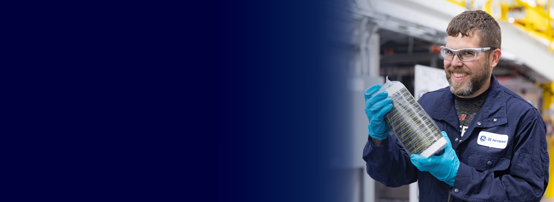 A GE Aerospace employee wearing blue gloves and a navy blue uniform holds a cylindrical component wrapped in protective material. The background features an industrial setting with machinery and equipment.
