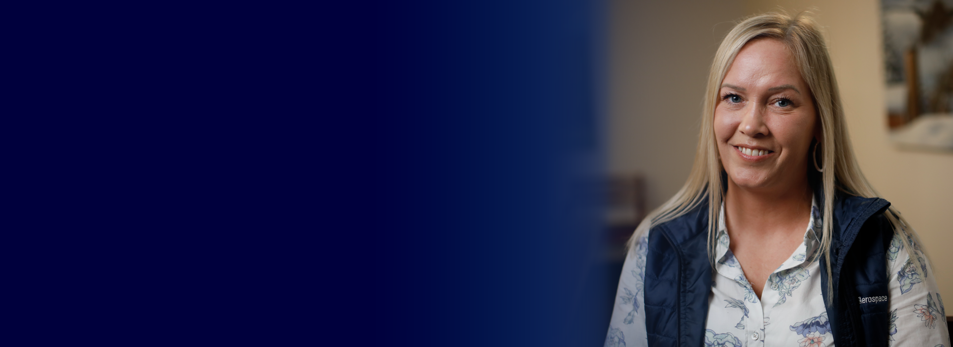 A person wearing a floral shirt and a navy vest with "GE Aerospace" embroidered on it, sitting indoors with a blurred background.