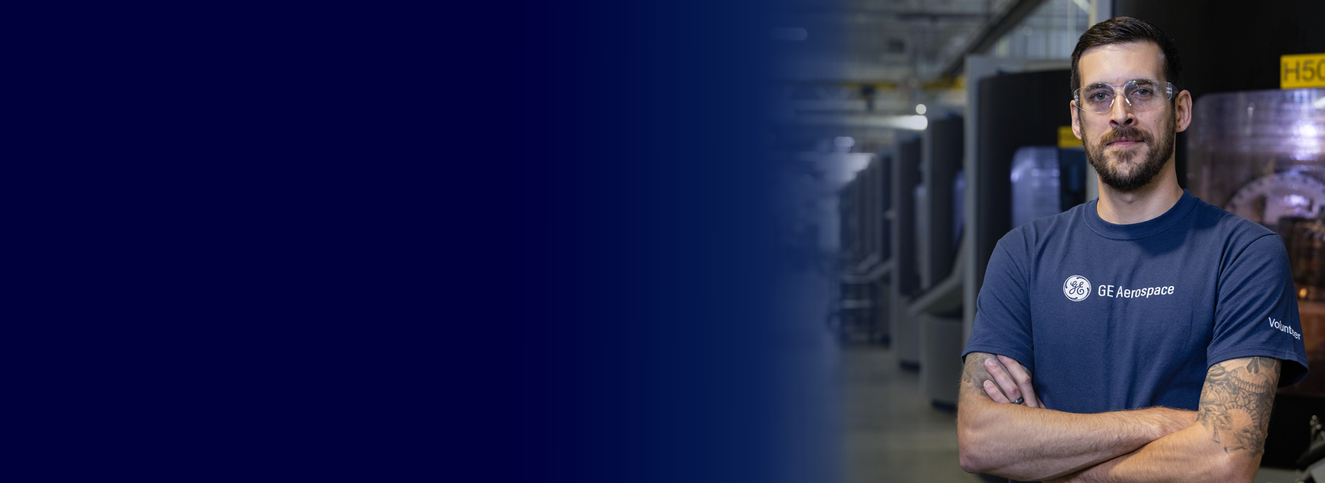 A person wearing a navy blue GE Aerospace t-shirt stands with arms crossed in an industrial setting. The background features machinery and equipment