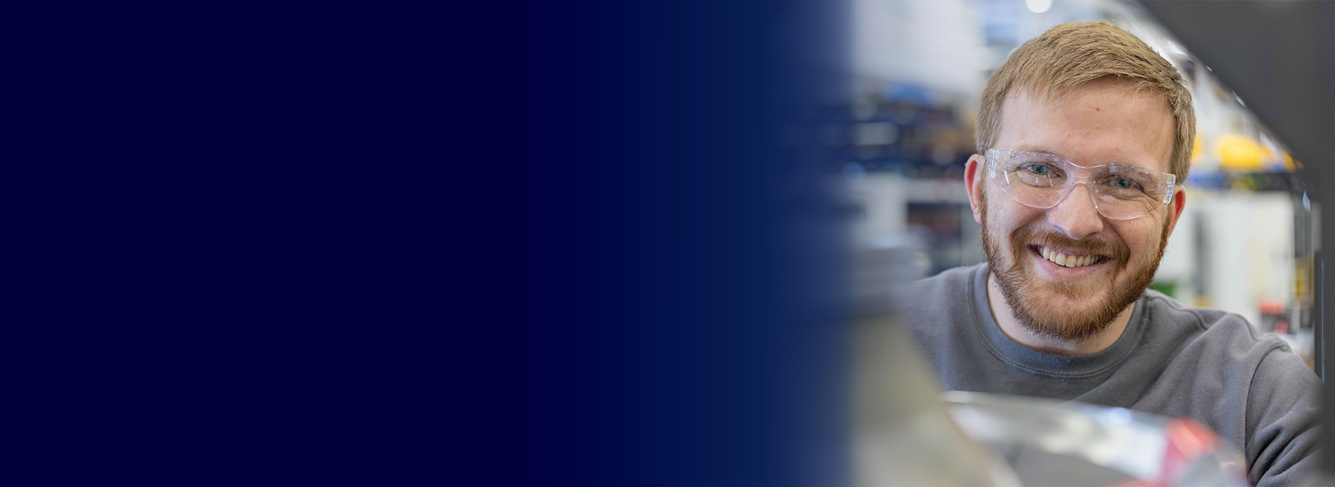 A person wearing a gray shirt is positioned in a workshop environment with blurred shelves and equipment in the background. The left side of the image features a solid dark blue gradient.
