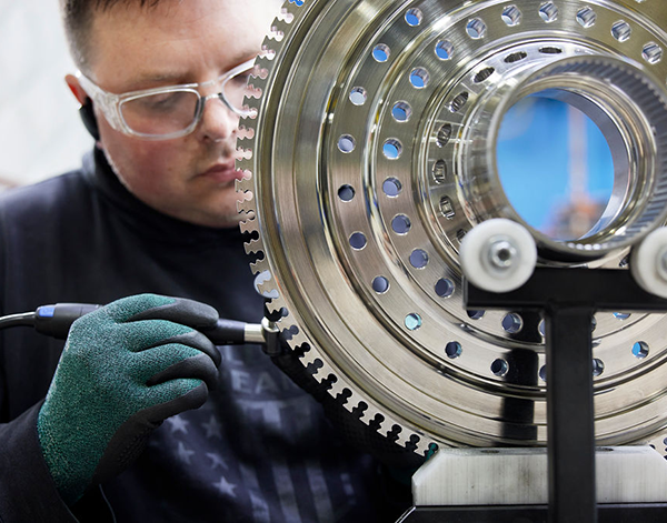 A worker wearing green gloves uses a tool to inspect or work on a metallic aerospace component with intricate holes and grooves.