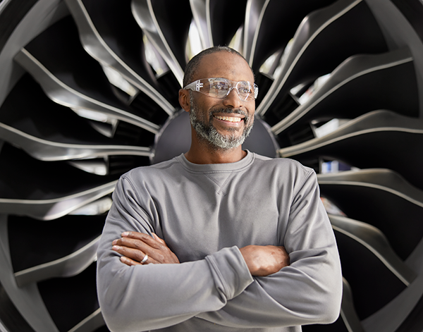 A person stands confidently with arms crossed in front of a large jet engine, showcasing the intricate design of the engine's fan blades. The individual is wearing a gray long-sleeve shirt, and the setting highlights the engineering and aerospace context.