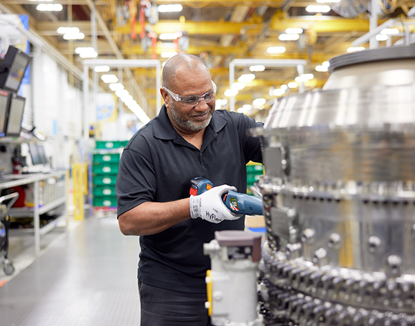 A worker in a manufacturing facility uses a power tool to perform maintenance on a large engine component. The workspace is brightly lit, with industrial equipment and storage areas visible in the background.