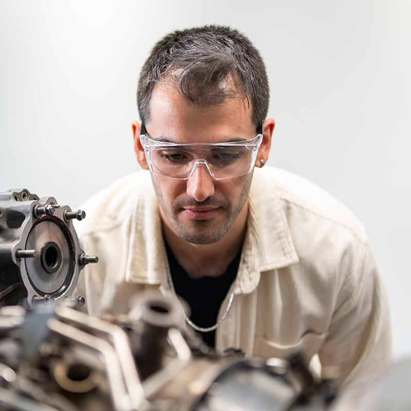 A person wearing safety glasses examines a complex mechanical component in a well-lit environment. The focus is on the machinery in the foreground.