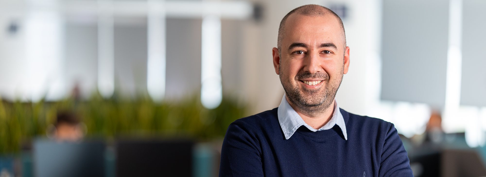 Man in a navy sweater with a light blue collared shirt stands with arms crossed in a modern, open-plan office; background shows soft-focus desks, colleagues, and indoor plants.