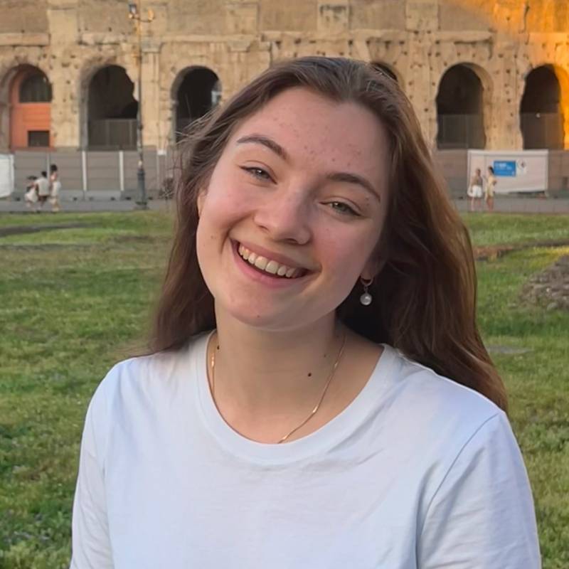A young woman with long brown hair smiles while standing outdoors in front of the Colosseum in Rome. She is wearing a white shirt and pearl earrings, with green grass and ancient stone arches in the background.