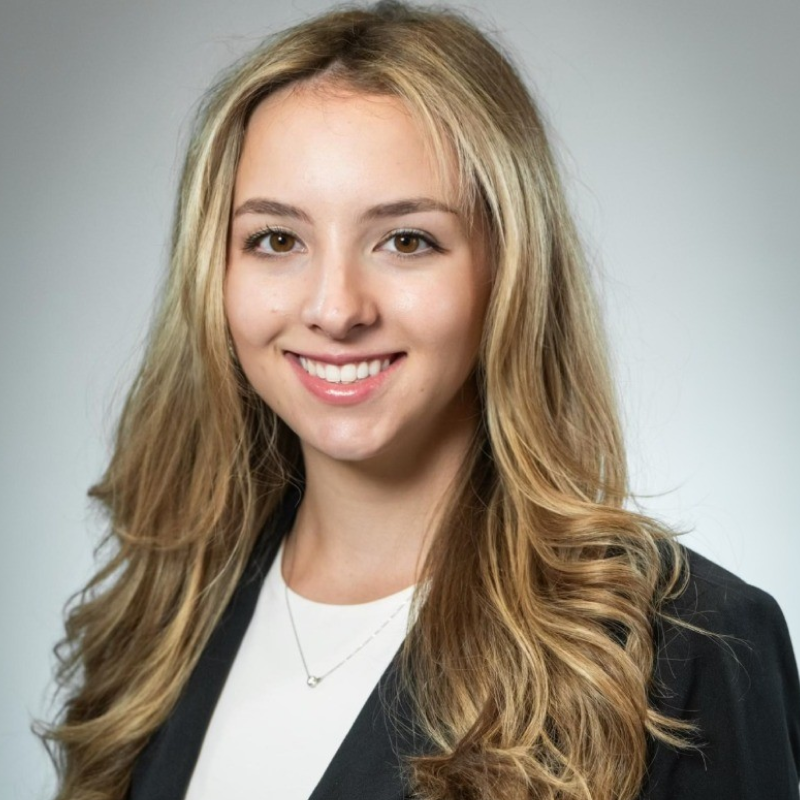 A young woman with long, wavy blonde hair smiles at the camera. She is wearing a black blazer over a white top and a small necklace, with a neutral, light gray background.