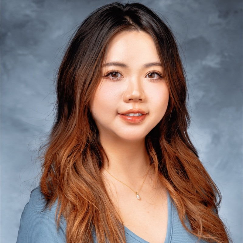 A young woman with long, wavy brown hair wearing a light blue top and a delicate necklace, smiling softly in front of a gray-blue studio background.