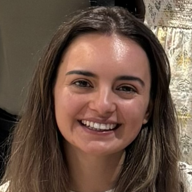A woman with long brown hair smiles at the camera; she is indoors and wearing a light-colored top, with a blurred background featuring another person and patterned fabric.