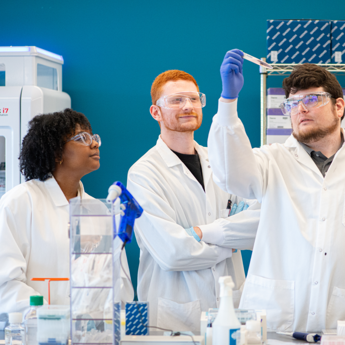 Three scientists wearing lab coats and safety glasses observe as one holds up a vial in a laboratory setting, with scientific equipment and supplies visible on the bench.