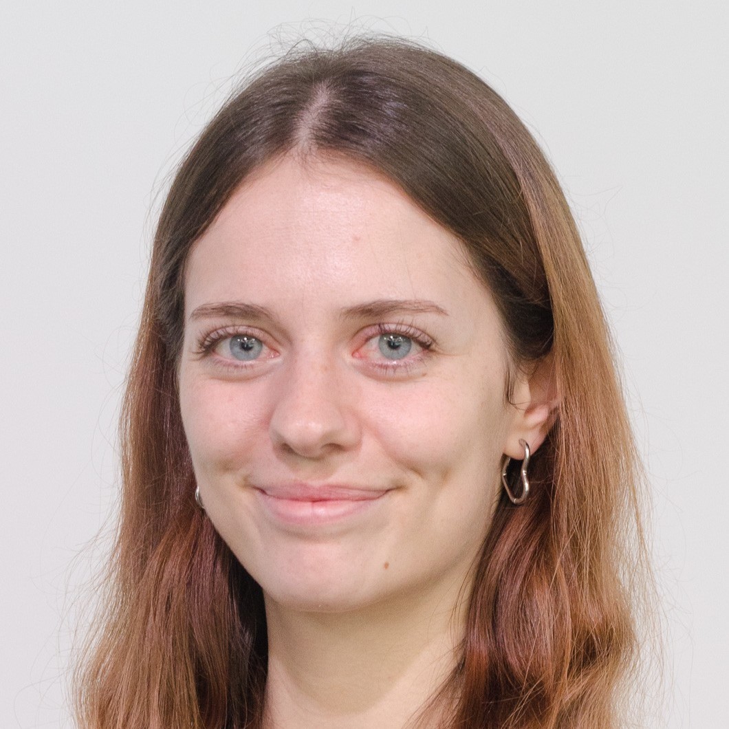 A young woman with long, wavy brown hair wearing a light blue top and a delicate necklace, smiling softly in front of a gray-blue studio background.