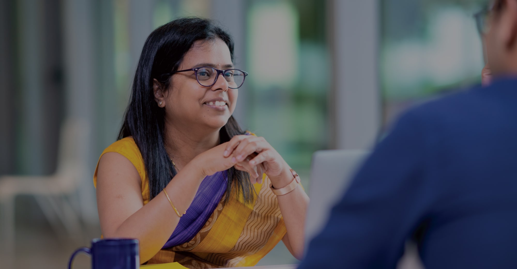 A smiling woman in a yellow saree with a purple border, sitting at a table and having a conversation with a colleague in a modern office setting. A blue coffee mug and a laptop are visible on the table.