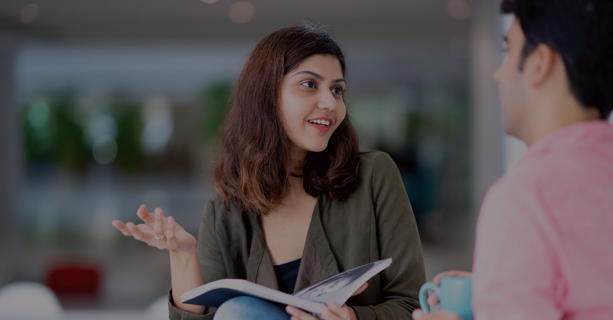 A smiling woman in conversation, holding a magazine while speaking to a colleague over coffee in a modern office setting.