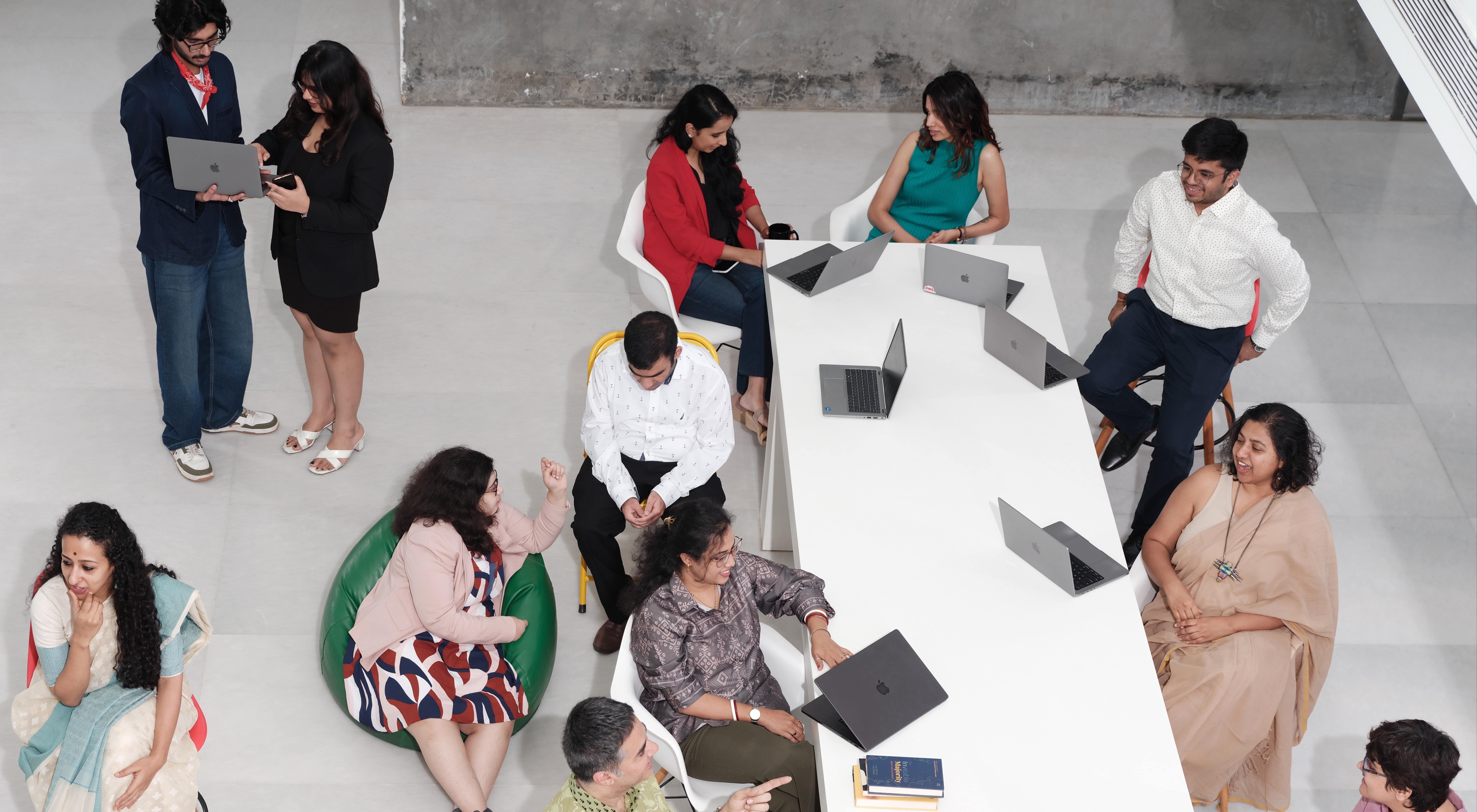 A diverse group of employees collaborating and socializing around a long white table with laptops in an open, inclusive workspace.