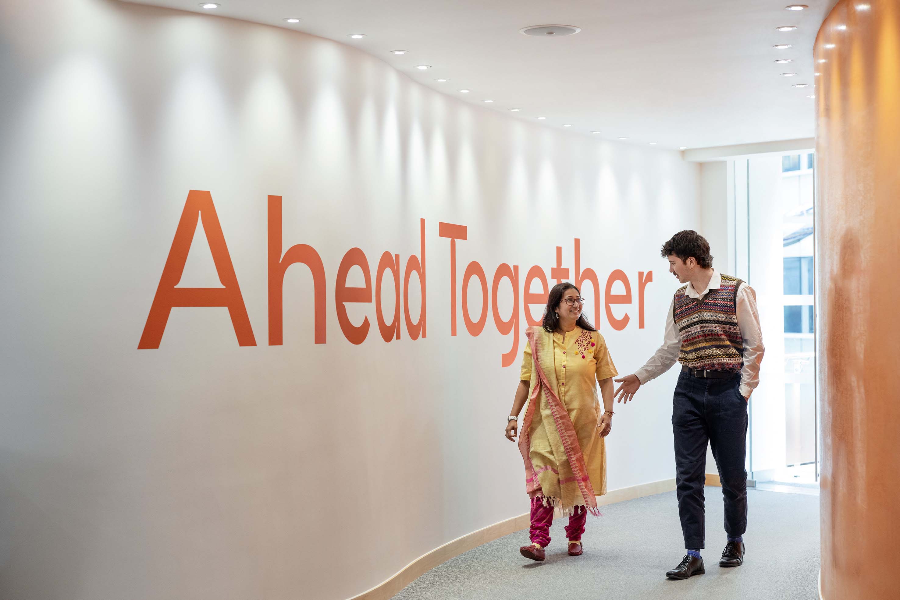 Two colleagues walk past a wall reading "Ahead Together" in a bright corridor as they talk.