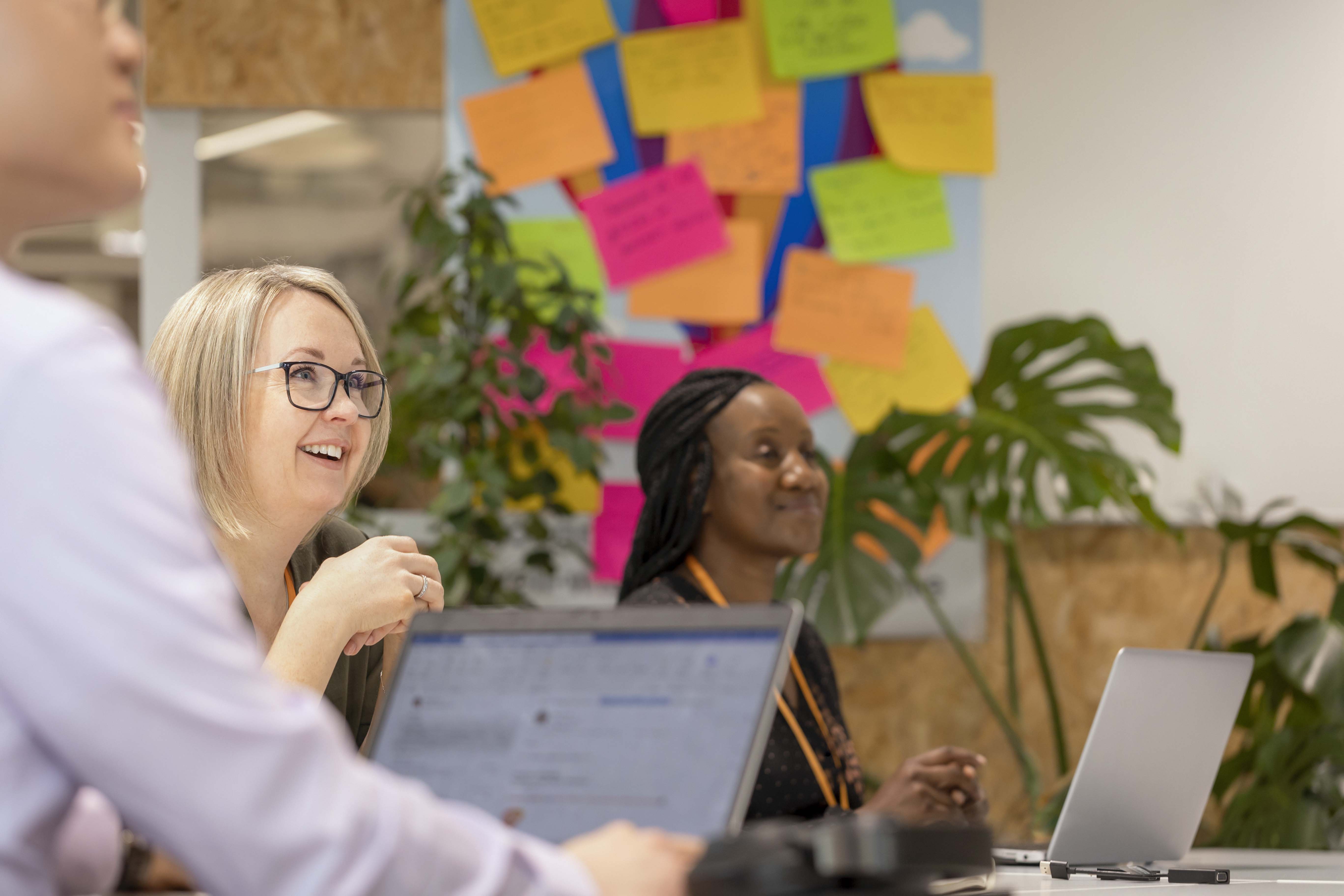Team meeting at a table with laptops; colorful sticky notes on the wall and plants add a creative backdrop.