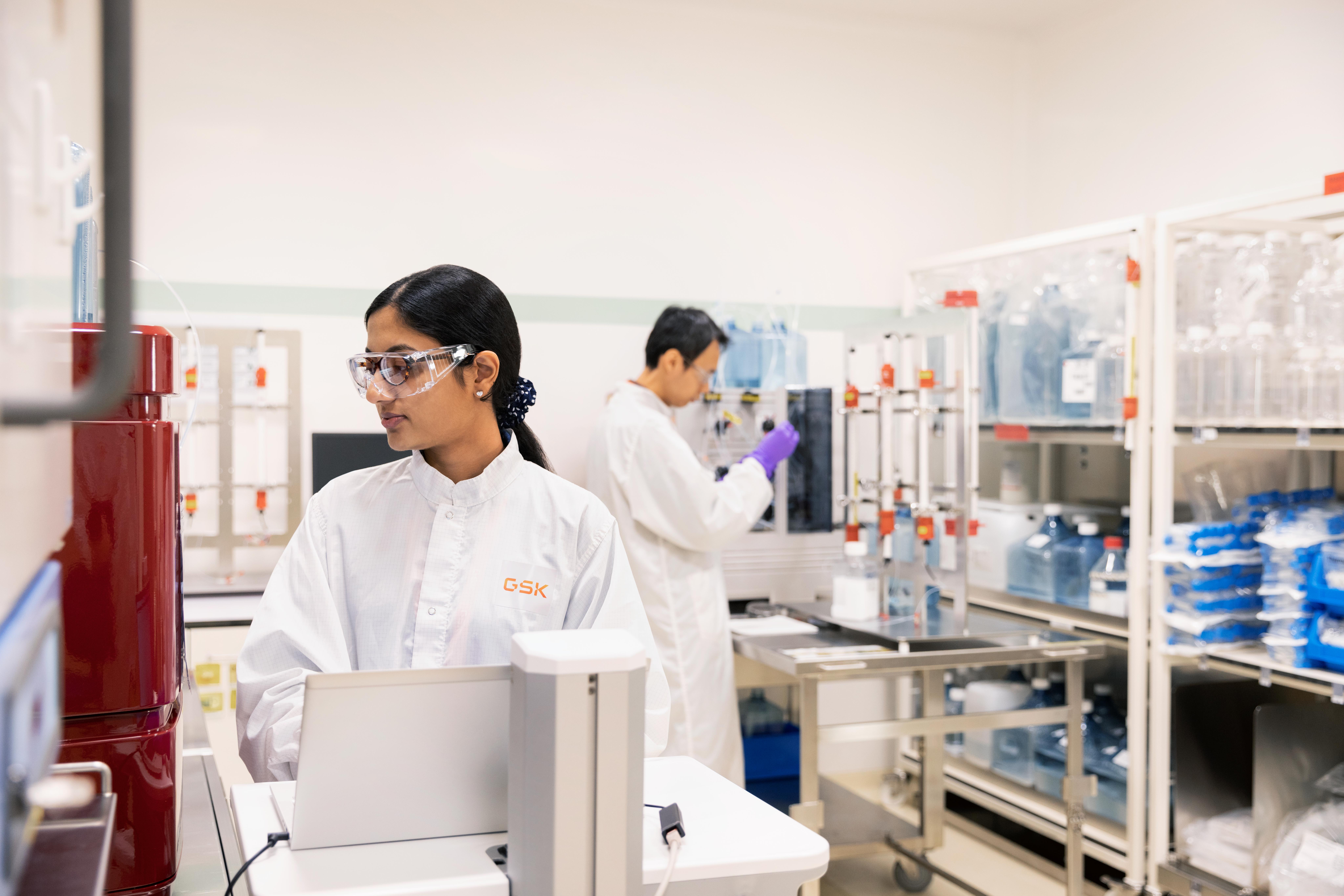 Two lab workers in white coats operating scientific equipment in a lab filled with machines and storage shelves.