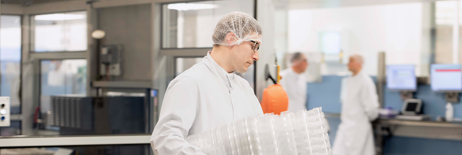 Worker in protective clothing and gloves carries a stack of clear plastic trays through a sterile production area.