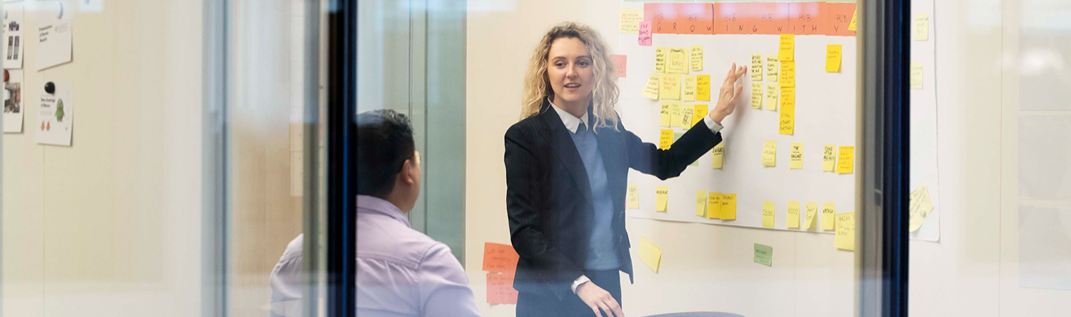 Small meeting room seen through glass; one person stands pointing at sticky notes on a wall while another sits and listens.