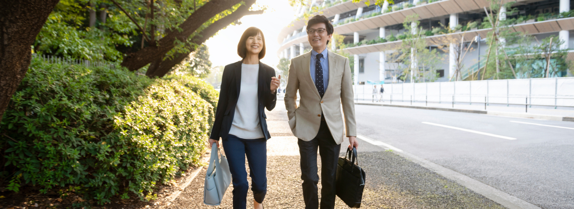 Two people in business attire walk and smile along a sunlit sidewalk bordered by greenery on one side and a modern building on the other, both carrying bags.
