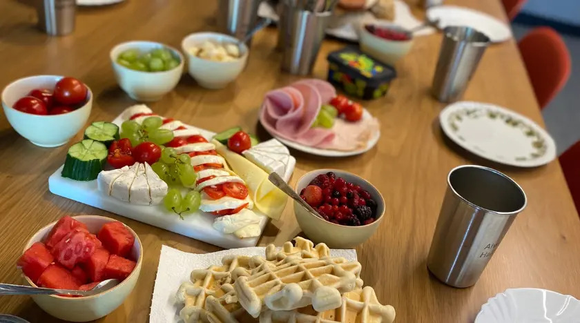 A wooden table set for breakfast with plates of cheese, cold cuts, fresh fruit, berries, cherry tomatoes, cucumbers, waffles, and metal cups. The food is neatly arranged and inviting.