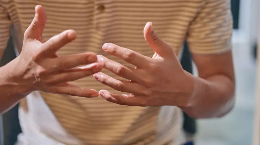 A person wearing a beige and white striped shirt gestures with both hands, fingers spread, as if explaining or emphasizing something. The image focuses on the hands and torso, with the head out of frame.