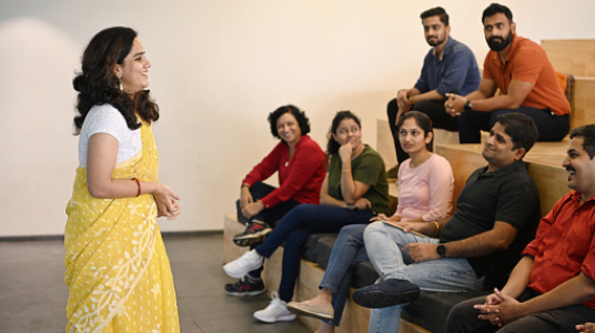 A woman in a yellow sari is speaking to a group of seven people, who are sitting on tiered benches and listening attentively in a modern, indoor setting.