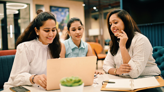 Three women sit together at a table, smiling and looking at a laptop screen. One has a notebook open in front of her, and there is a small potted plant on the table. The setting appears to be a modern office or workspace.