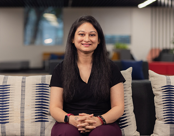 A woman with long dark hair, wearing a black shirt and maroon pants, sits on a patterned sofa, smiling at the camera in a modern office setting.